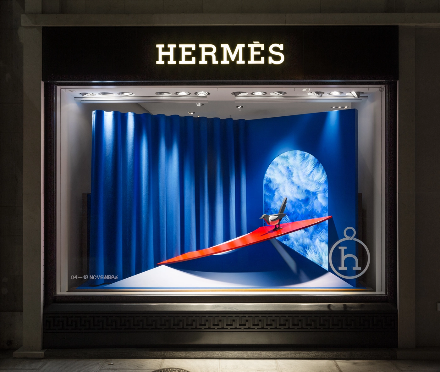 Hermès retail window display featuring a blue background, screen, and stylized bird perched on a red plate